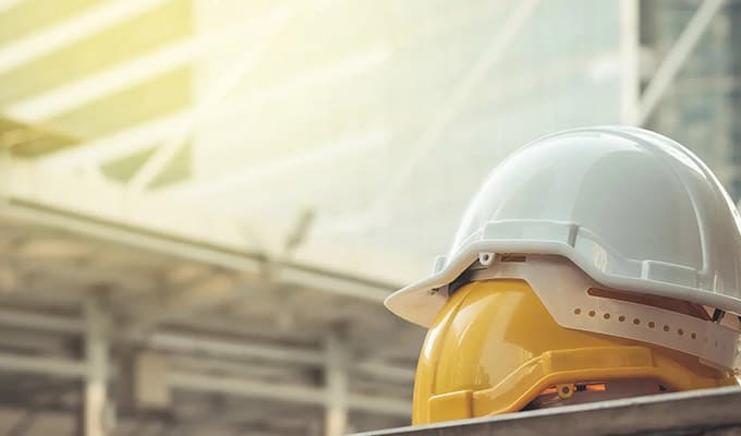 Construction scene looking out onto a bridge with strong sunshine, two construction helmets stacked on top of each other, one helmet is yellow, the other construction helmet is white