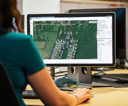 A person sits at a desk looking at a computer monitor displaying a satellite map with marked locations, using MicroStation to plan new campuses.